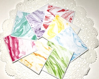 Op de afbeelding: Acht vierkante kaarten met marmerpatronen in verschillende kleuren, waaronder groen, paars, rood, geel, blauw en wit. De kaarten zijn in een cirkelvormige opstelling op een witte kantklos geplaatst.
