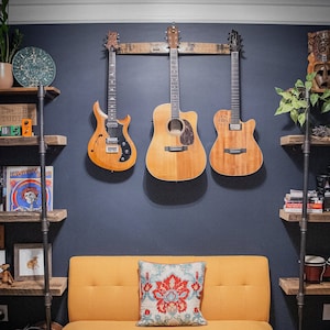 Puede incluir: Una habitación con un sofá color mostaza y un cojín decorativo con diseño floral. Tres guitarras cuelgan de la pared sobre el sofá. Estanterías de madera con decoración y plantas a ambos lados del sofá.