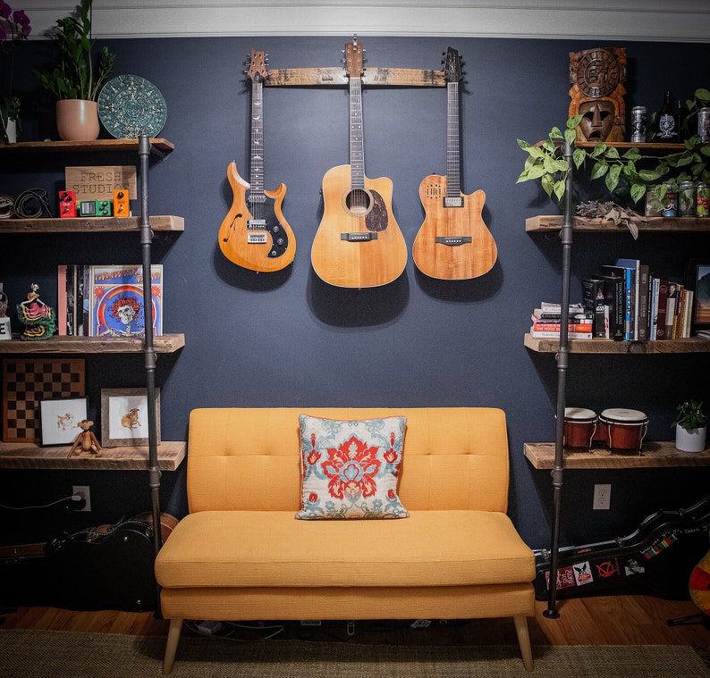 Handcrafted whiskey barrel stave guitar hanger displaying three guitars on a dark blue wall above a modern mustard yellow couch, with eclectic room decor and bookshelves in a cozy music-themed living space.