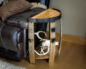 May include: A rustic, half-circle side table made from reclaimed wood and metal barrel hoops. The table features a natural wood finish with black accents and a lower shelf. Antlers are displayed on the shelf. The table is next to a brown leather recliner.