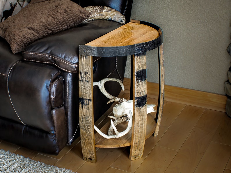 May include: A rustic, half-circle side table made from reclaimed wood and metal barrel hoops. The table features a natural wood finish with black accents and a lower shelf. Antlers are displayed on the shelf. The table is next to a brown leather recliner.
