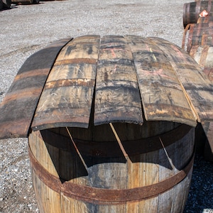 May include: Several curved wooden barrel staves with weathered, dark brown and charcoal-colored surfaces are arranged horizontally on top of a large, upright wooden barrel. The staves show signs of age, including visible wood grain and texture.