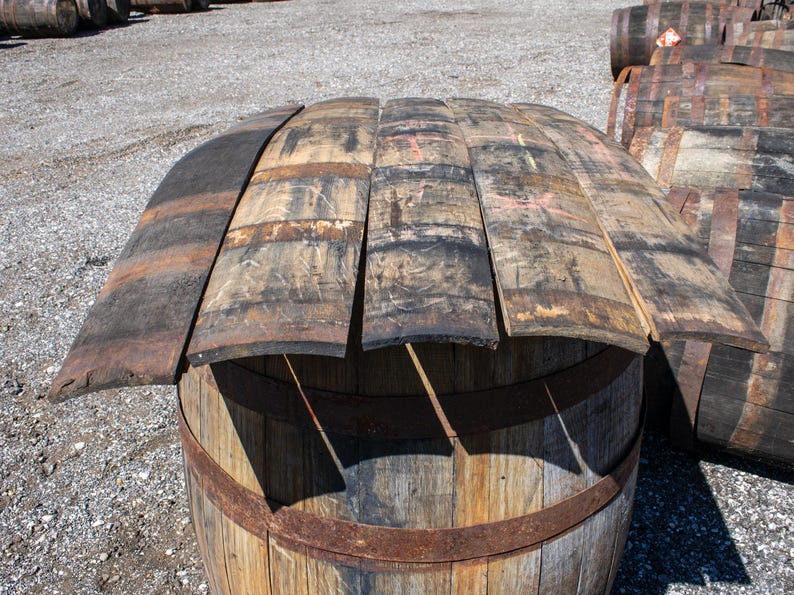 May include: Several curved wooden barrel staves with weathered, dark brown and charcoal-colored surfaces are arranged horizontally on top of a large, upright wooden barrel. The staves show signs of age, including visible wood grain and texture.