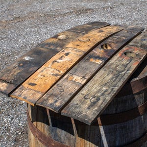 May include: Four reclaimed wooden barrel staves with weathered brown and charcoal gray tones, featuring visible circular bung holes, natural wood grain textures, and faint black ink markings, resting horizontally atop a cylindrical wooden barrel.