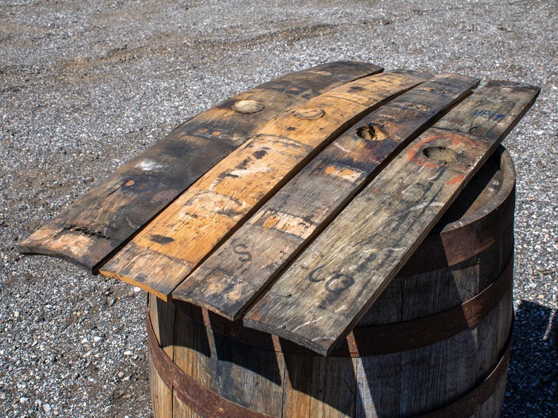 May include: Four reclaimed wooden barrel staves with weathered brown and charcoal gray tones, featuring visible circular bung holes, natural wood grain textures, and faint black ink markings, resting horizontally atop a cylindrical wooden barrel.