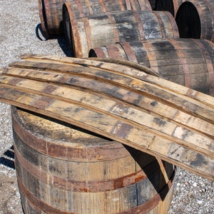 May include: A collection of curved wooden barrel staves with weathered, dark brown grain patterns and charred edges, resting on top of a large, cylindrical wooden barrel with rusted metal hoops, surrounded by several other similar barrels.