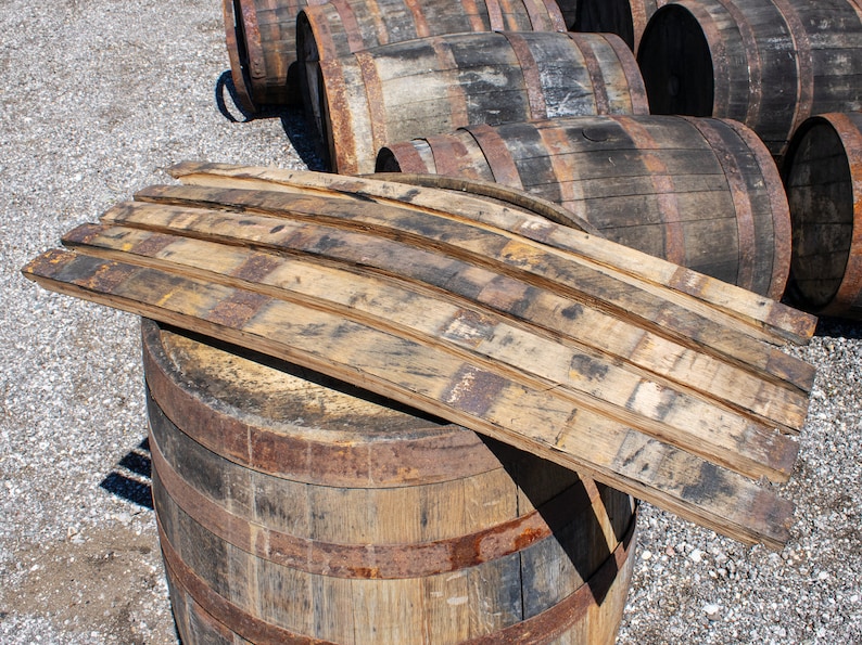 May include: A collection of curved wooden barrel staves with weathered, dark brown grain patterns and charred edges, resting on top of a large, cylindrical wooden barrel with rusted metal hoops, surrounded by several other similar barrels.