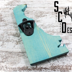 May include: A teal blue wooden bottle opener shaped like the state of Delaware. The bottle opener has a black metal opener attached to it. The background is a whitewashed wood surface. The text "Skull Creek Designs" is visible in the upper right corner of the image.