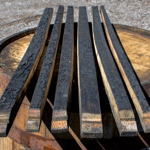 May include: Five long, narrow wooden barrel staves with a heavily charred, black, alligator-skin texture on the top surface. The wood grain is visible on the light brown, uncharred sides and ends of each stave, which are arranged in a parallel row.