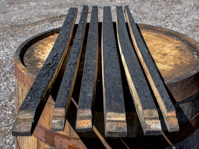 May include: Five long, narrow wooden barrel staves with a heavily charred, black, alligator-skin texture on the top surface. The wood grain is visible on the light brown, uncharred sides and ends of each stave, which are arranged in a parallel row.
