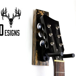 May include: A black electric guitar with a wooden wall mount. The guitar has a black body and a black headstock with silver tuning pegs. The wall mount is made of reclaimed wood and has a distressed finish.