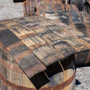 May include: A collection of weathered, curved wooden barrel staves arranged in a stack on top of a large, cylindrical wooden barrel. The staves feature dark, charred wood grain patterns, rusted metal bands, and visible signs of aging and wear.