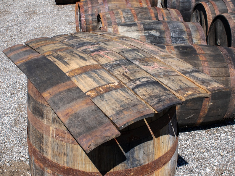 May include: A collection of weathered, curved wooden barrel staves arranged in a stack on top of a large, cylindrical wooden barrel. The staves feature dark, charred wood grain patterns, rusted metal bands, and visible signs of aging and wear.