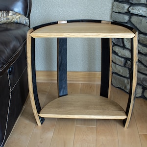 May include: A handcrafted side table made from reclaimed oak barrel staves. The table features a light oak top and shelf, supported by curved black bands. The design incorporates the natural curvature of the barrel, creating a unique, rustic aesthetic.
