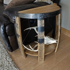 May include: Half-circle wooden side table made from a repurposed barrel. The table features a dark metal band around the top and a lower shelf. Inside, there are deer antlers and a skull. The table is next to a brown leather couch.