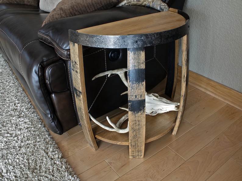 May include: Half-circle wooden side table made from a repurposed barrel. The table features a dark metal band around the top and a lower shelf. Inside, there are deer antlers and a skull. The table is next to a brown leather couch.