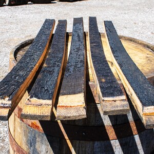May include: Five long, curved wooden barrel staves arranged in a row. Each stave features a deeply charred, black textured surface on one side, contrasting with the natural light brown wood grain visible on the edges and the bottom ends.