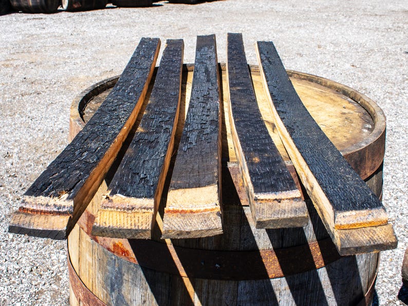 May include: Five long, curved wooden barrel staves arranged in a row. Each stave features a deeply charred, black textured surface on one side, contrasting with the natural light brown wood grain visible on the edges and the bottom ends.