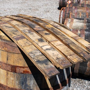 May include: Several curved wooden barrel staves with a weathered, light brown finish and dark charred markings are stacked on top of a large, cylindrical wooden barrel featuring rusted metal hoops and a natural wood grain texture.