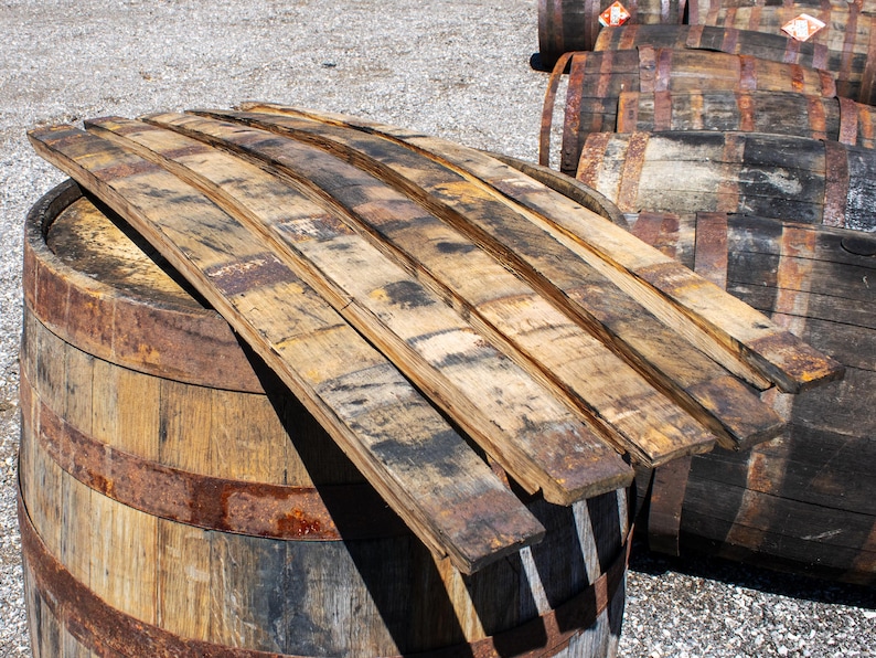May include: Several curved wooden barrel staves with a weathered, light brown finish and dark charred markings are stacked on top of a large, cylindrical wooden barrel featuring rusted metal hoops and a natural wood grain texture.