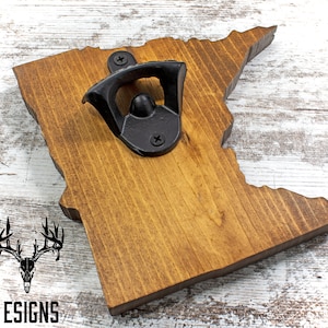 May include: A wooden bottle opener shaped like the state of Minnesota. The opener is stained a dark brown color and has a black metal bottle opener attached to it. The opener is sitting on a white wooden surface.