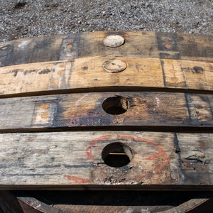 May include: Four weathered wooden barrel staves arranged horizontally. The two center staves feature circular holes, while the outer staves have circular wooden plugs. The wood displays a natural grain, dark charred sections, and faint markings.