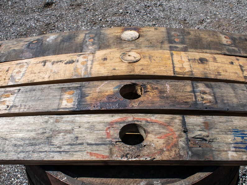 May include: Four weathered wooden barrel staves arranged horizontally. The two center staves feature circular holes, while the outer staves have circular wooden plugs. The wood displays a natural grain, dark charred sections, and faint markings.