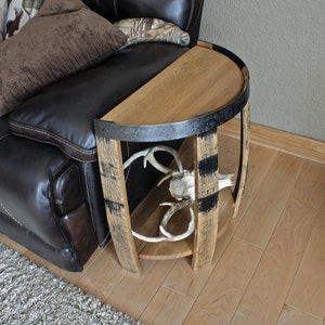 May include: A rustic side table crafted from a repurposed barrel. The table features a half-circle tabletop with a metal band and a lower shelf. Inside, a deer skull and antlers are displayed. The table is next to a brown leather couch.