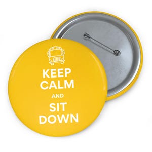 Puede incluir: Un botón amarillo con el texto "KEEP CALM AND SIT DOWN" en blanco, junto con un gráfico blanco de un autobús escolar. El botón tiene un alfiler plateado en la parte posterior.