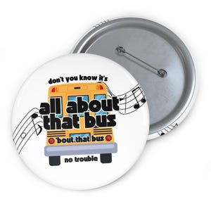 Puede incluir: Un botón blanco con un diseño de autobús escolar amarillo y el texto "don't you know it's all about that bus" con notas musicales. El botón tiene una parte trasera de metal y un alfiler.