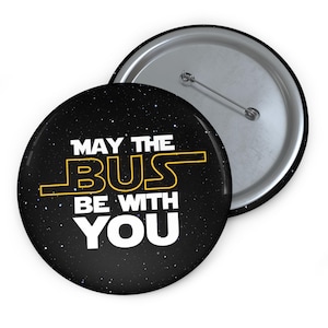 Puede incluir: Un botón redondo negro con el texto "MAY THE BUS BE WITH YOU" en blanco y amarillo. El botón tiene un reverso plateado con un alfiler y un fondo estrellado.
