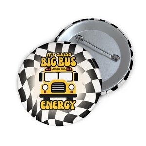 Puede incluir: Dos botones redondos con un patrón de cuadros blancos y negros. Un botón presenta un gráfico de autobús escolar amarillo con el texto "IT'S GIVING BIG BUS ENERGY SCHOOL BUS". El otro botón es plateado.