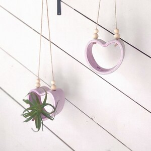 May include: Two pink heart-shaped hanging planters with wooden beads. The planter on the left has a small green plant inside. Both planters are hanging from black metal hooks.
