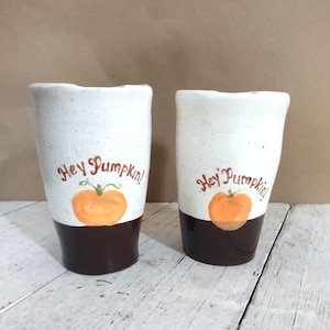 May include: Two ceramic tumblers with a white speckled upper half and a dark brown base. Each tumbler features the phrase "Hey Pumpkin!" and a hand-painted orange pumpkin design, perfect for fall.