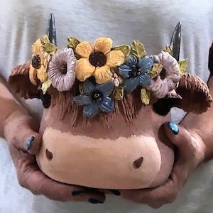 May include: A brown ceramic cow planter with a floral crown. The crown is made of yellow, blue, and pink flowers.