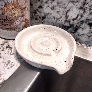 May include: A round, off-white ceramic sponge holder featuring a speckled pattern with dark brown dots. The interior surface has a raised circular ridge design and a small spout at the front edge to allow for drainage, providing a functional base.