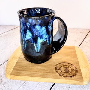 May include: A ceramic mug with a glossy black base and a vibrant blue drip glaze pattern cascading from the rim. The mug features a wide, curved handle and sits on a rectangular bamboo board engraved with a Michigan state map and text.