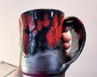 Może przedstawiać: Ręcznie robiony ceramiczny kubek z unikalnym szkliwem. Kubek ma czarną podstawę, która przechodzi w srebrno-szarą środkową część, oraz efektowny czerwono-czarny wzór na górnej części. Duży uchwyt jest czarny i błyszczący.