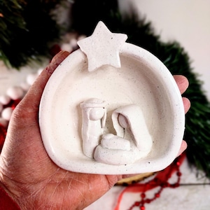 May include: White ceramic nativity scene ornament with a star on top. The scene depicts Mary, Joseph, and baby Jesus in a manger.