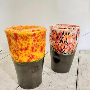 May include: Two ceramic tumblers. One is yellow with red and green speckles, the other has a white top with orange and red floral patterns. Both have dark gray bases and are on a light wood surface.