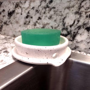May include: A white ceramic soap dish with black speckles holds a green bar of soap. The dish has a small hole on the side and sits on the edge of a stainless steel sink. The background is a speckled granite countertop.