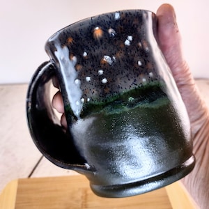 Może przedstawiać: Ręcznie robiony ceramiczny kubek z ciemnym, nakrapianym szkliwem. Kubek ma czarną podstawę, która przechodzi w głęboką zieleń, a następnie w nakrapiany, ciemnoniebiesko-czarny brzeg. Uchwyt jest czarny. Unikalny element do kawy lub herbaty.