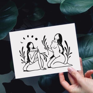 May include: Black and white line art print of two figures with long hair, standing in a field of flowers. The figures are nude and have their arms raised. The print has a minimalist style.