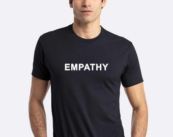 May include: Black t-shirt with the word "EMPATHY" printed in white block letters. The shirt is a crew neck style and appears to be made of a soft material. The model is wearing a silver bracelet.