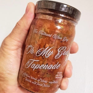 May include: A glass jar filled with a red and green tapenade, labeled "The Tattooed Wine Guy Oh My God Tapenade". A delicious blend of olives, vegetables, spices, and fresh juices.