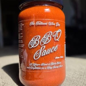 May include: A jar of BBQ sauce with a dark lid. The label reads "The Tattooed Wine Guy BBQ Sauce" in white script. The sauce is a deep red color, and the label states it's a unique blend of spices, juices, and vegetables.