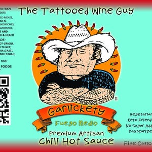 Könnte beinhalten: Ein rotes, weißes und blaues Etikett mit einer Cartoon-Illustration eines tätowierten Mannes, der einen Cowboyhut trägt. Das Etikett ist für eine Chili-Sauce namens "Garlickety Fuego Medio Premium Artisan Chili Hot Sauce". Das Etikett enthält auch einen QR-Code, den Text "The Tattooed Wine Guy Foods" und eine Liste von Weinpaarungen.