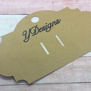 Puede incluir: Una tarjeta de exhibición de aretes marrón con una forma decorativa y un agujero en la parte superior. La tarjeta tiene dos ranuras para aretes y el texto "yDesigns" en escritura negra.