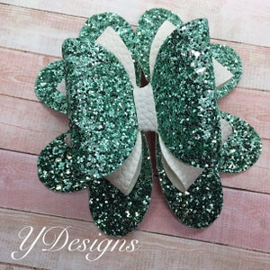 May include: A large, green glitter bow with white accents. The bow is made of multiple layers of fabric and has a unique, layered design.