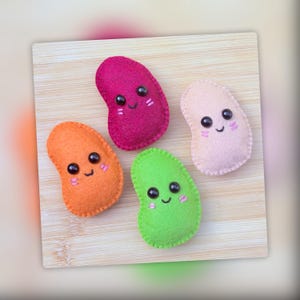 May include: Four felt bean-shaped plush toys in bright colors including magenta, orange, lime green, and pale pink. Each plush features two black plastic bead eyes, a stitched black smile, and two small pink stitched lines on each cheek.